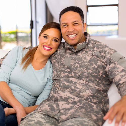 A couple sit close together on a sofa, smiling. One wears a military uniform and the other rests against his shoulder.