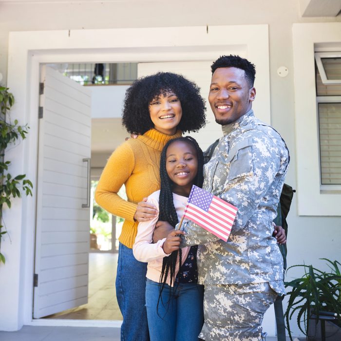 A service member in uniform stands at the front door of a home with his wife and daughter, who holds a small American flag. All three are smiling.